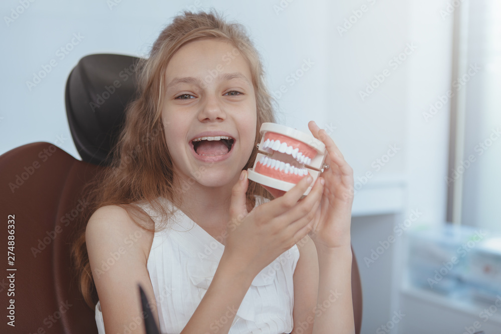 Happy cute young girl having fun, playing with dentures, laughing to the camera, sitting in a dental chair. Lovely little patient on medical dental appointment, copy space. Healthy teeth concept Happy cute young girl having fun, playing with dentures, laughing to the camera, sitting in a dental chair. Lovely little patient on medical dental appointment, copy space. Healthy teeth concept