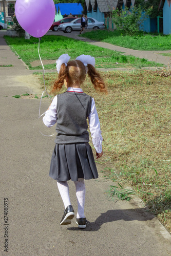 A girl with large white bows on her head, a beautiful hairdo in a school uniform, is walking along the sidewalk from the school. She is wearing a school uniform. View from the back