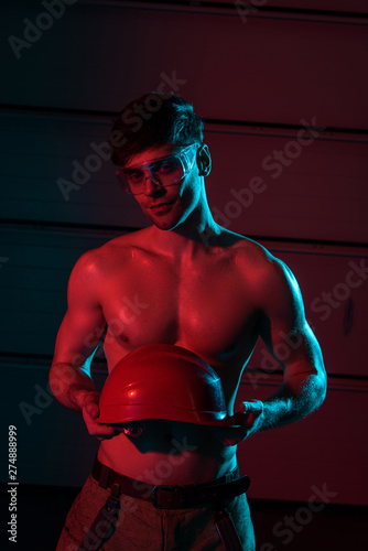 sexy shirtless fireman in protective goggles holding hardhat in darkness