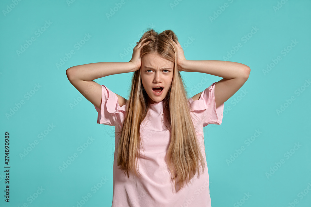 Studio portrait of a beautiful girl blonde teenager in a pink t-shirt posing over a blue background.