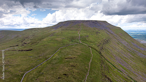 A panoramic aerial view of a mountain rocky summit with grassy green slope and trail path under a majestic blue sky and white clouds photo