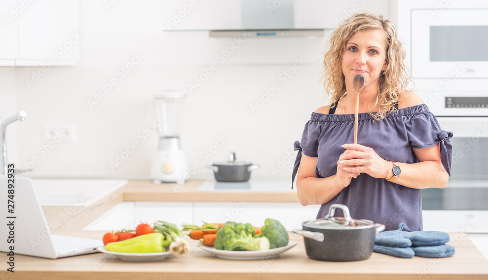Portrait of happy adult woman in her modern kitchen with pot and vegetables