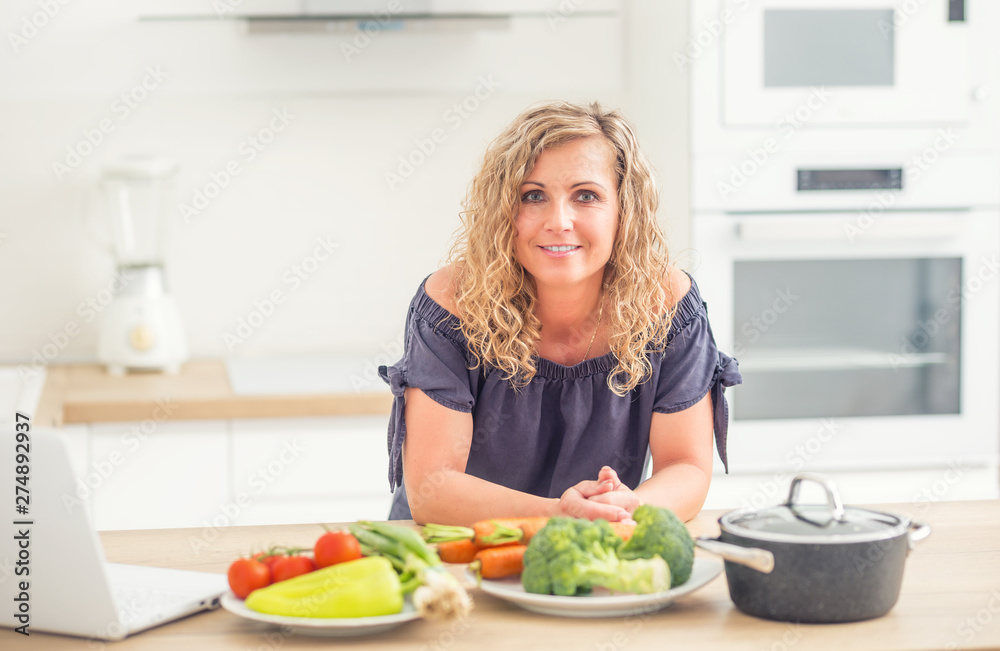 Portrait of happy adult woman in her modern kitchen with pot and vegetables