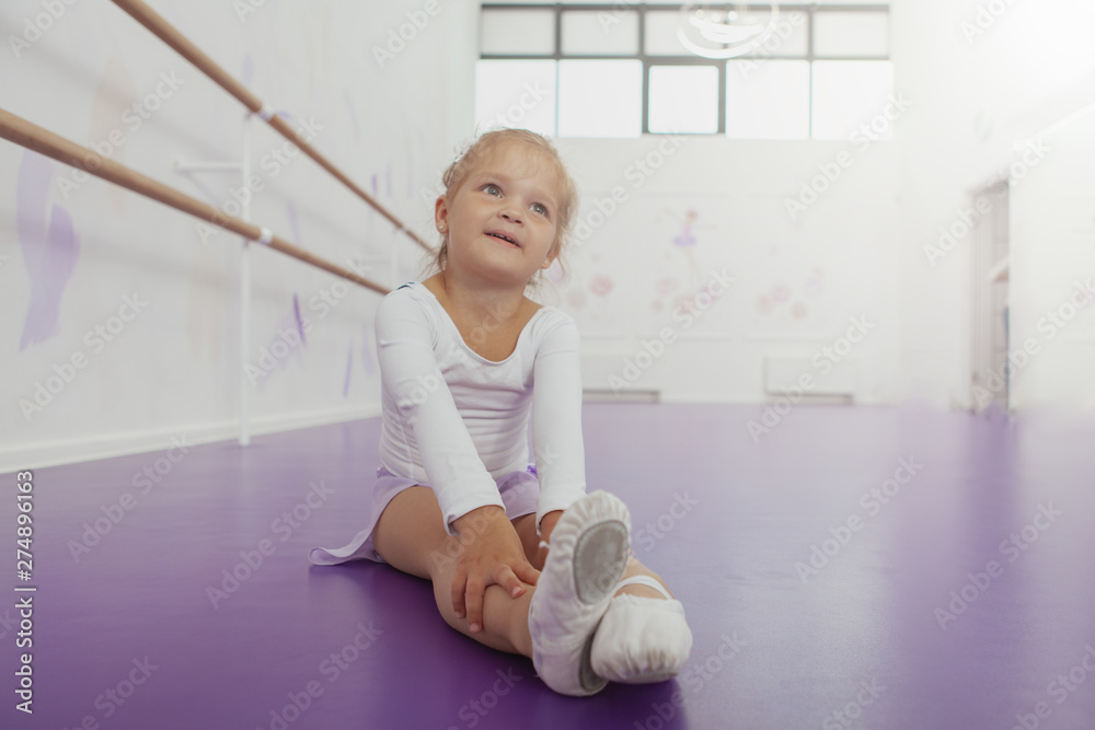 Charming little ballerina sitting on the floor at ballet class, looking away dreamily. Cute little girl in leotard, resting after exercising at ballet school, copy space