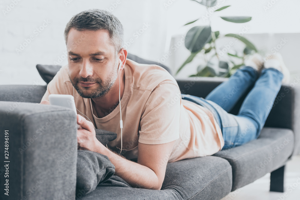 smiling man listening music in earphones and using smartphone while resting on sofa