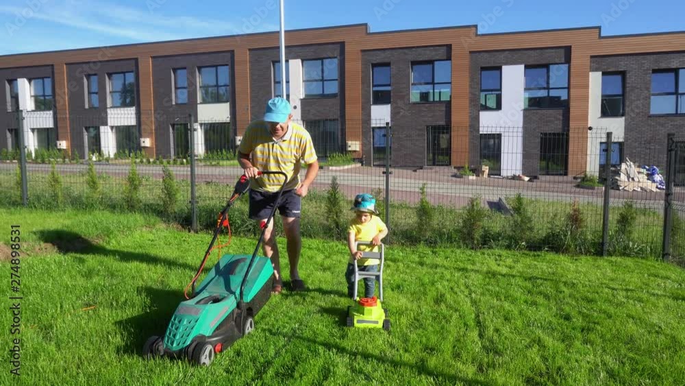 Small boy mows grass just like his dad. Father and son care lawn. Gimbal shot Small boy mows grass just like his dad. Father and son care lawn. Gimbal shot