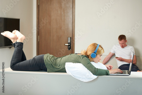 Pensive young woman in headphones programming on laptop whne her boyfriend texting friend in background photo