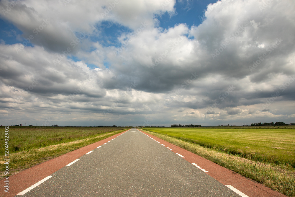 Road in Holland with red cycle path on both sides, perspective, under heavy dark threatening cloudy skies and between green meadows and a faraway horizon.