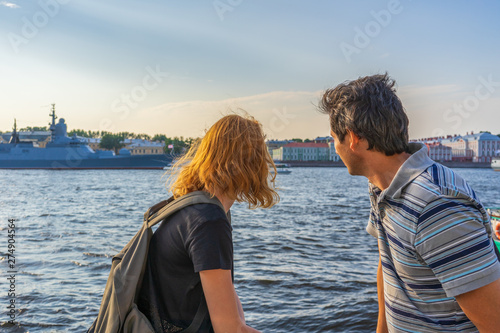 Middle-aged dark-haired man and young redhead lady on Neva river embankment looking at warships and architectural ensemble in summer evening at sunset. Travel concept. Saint Petersburg, Russia