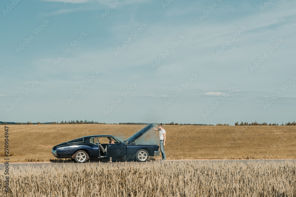 Obraz premium Couple standing near overheated car in the field, bright sunlight, steam under the hood