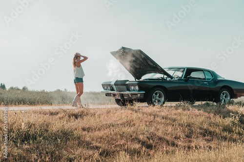 Young female standing near overheated car in the field, bright sunlight, steam under the hood