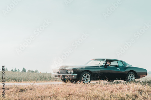 Young female sits in the overheated car in the field, bright sunlight, steam under the hood