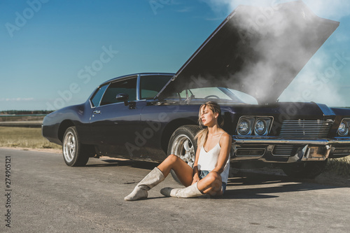 Young female sits near overheated car in the field, bright sunlight, steam under the hood photo