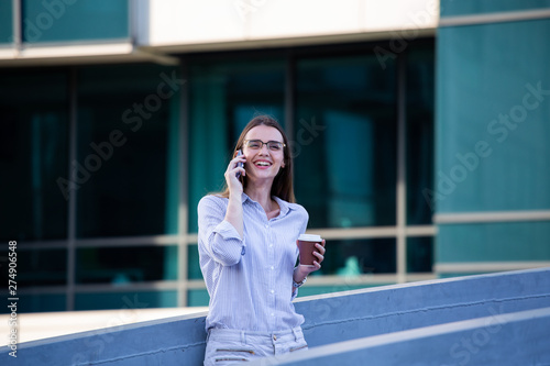Executive business woman looking at mobile smartphone and drinking coffee from disposable paper cup in the street with office buildings in the background