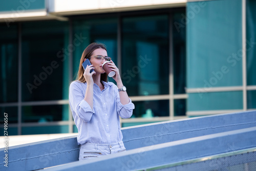 Executive business woman talking on mobile smartphone and drinking coffee from disposable paper cup in the street with office buildings in the background