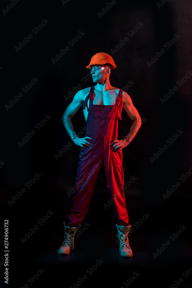 full length view of sexy fireman in overall, hardhat and protective goggles with arms akimbo on black