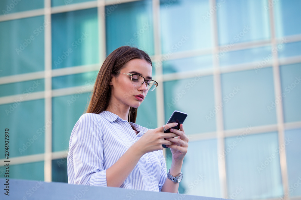 Executive business woman looking at mobile smartphone in the street with office buildings in the background Executive business woman looking at mobile smartphone in the street with office buildings in the background