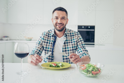 Portrait of charming imposing mature guy sit table hold hand fork knife want cut pleasant evening cheerful checked shirt big light apartment wineglass