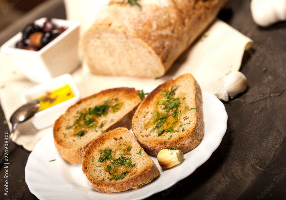 Sliced traditional toast bread with olive oil, olives, garlic and herbs on a plate and dark table