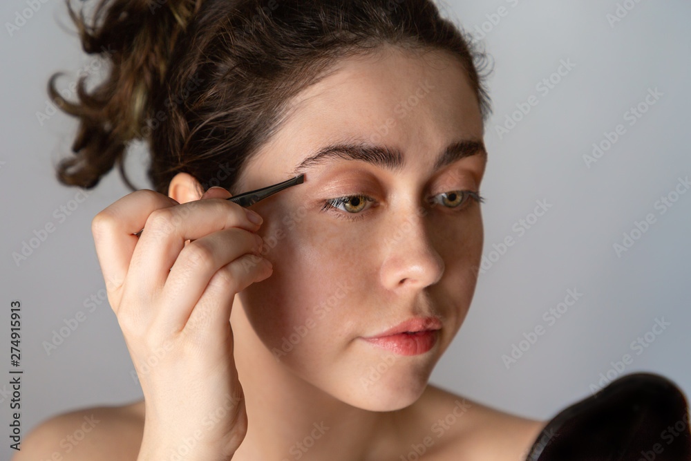 A young woman looks in the mirror and plucks her eyebrows. The concept of getting rid of unwanted facial hair A young woman looks in the mirror and plucks her eyebrows. The concept of getting rid of unwanted facial hair