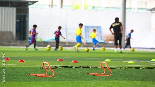 orange hurdles on green artificial turf with blurry coach and kid soccer are training. kid soccer players are jogging and control ball. soccer equipment in football academy.