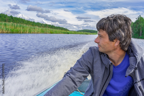 Handsome middle-aged man sitting at boat stern, floating along northern river and relaxing on beautiful landscape background in summer day. Travel concept. Chernaya river, Karelia, Russia