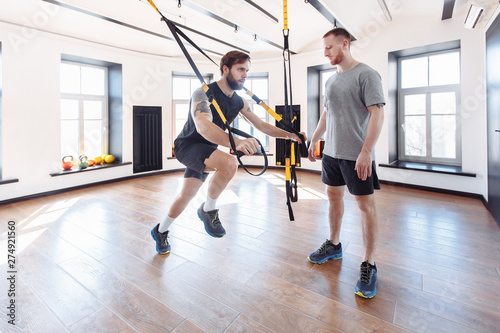 Male trainer teaches his student how to workout with suspended training system. Concept of strength training and work with the support of a coach
