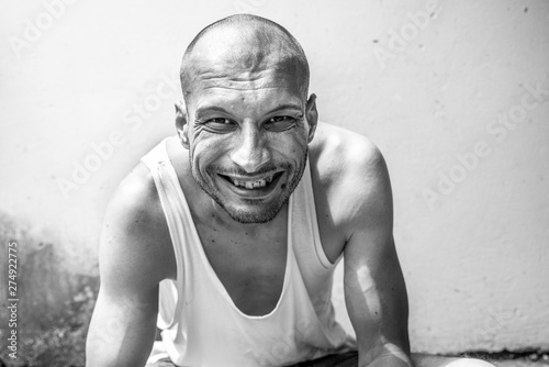 Young positive skinny anorexic bald happy smiling homeless man sitting on the urban street in the city or town near wall with big smile looking at the camera, homelessness social documentary concept photo