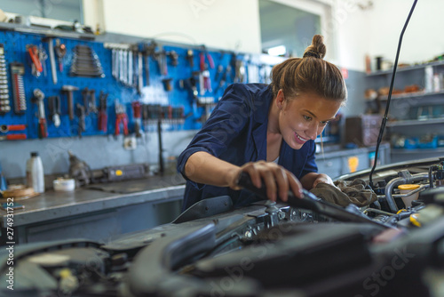 Lovely female auto mechanic smiling, examining engine of an automobile. Cheerful female car technician enjoying working at the garage, copy space. Repair, car service concept