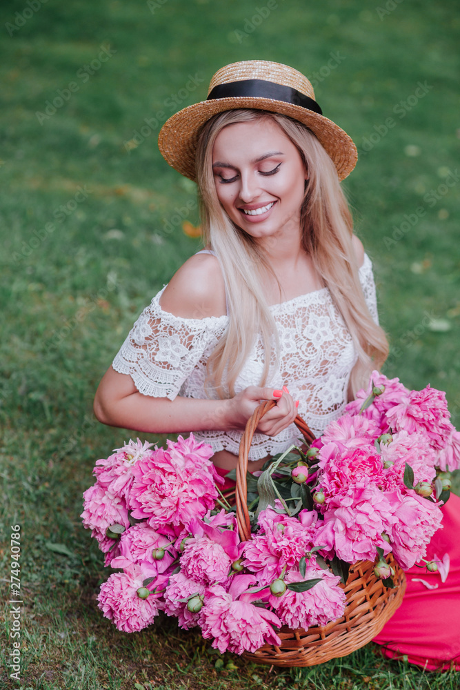 Fototapeta premium Beautiful girl in vintage dress and straw hat sitting on grass and holding pink peonies in rustic basket
