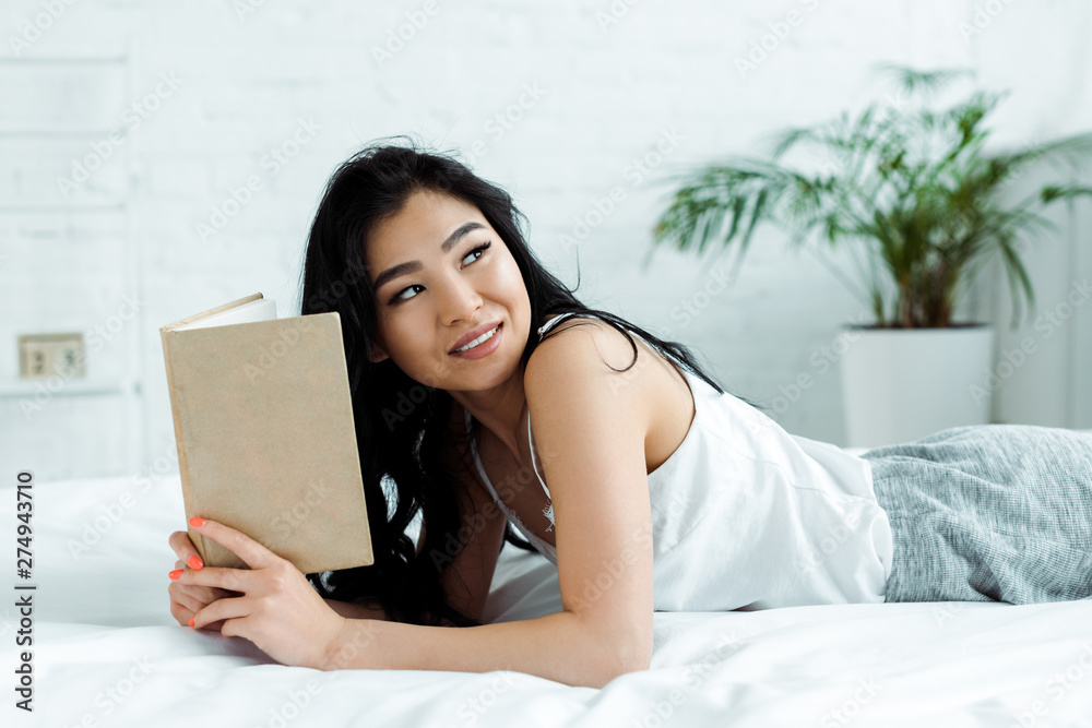 positive asian girl holding book while lying on bed at home positive asian girl holding book while lying on bed at home