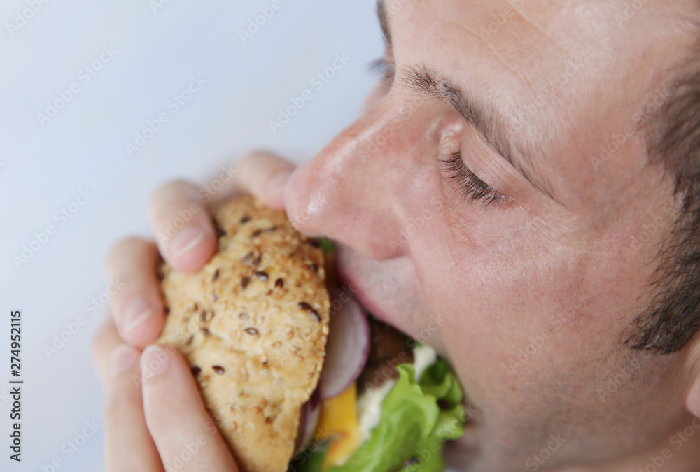 Close-up. A hungry young man eats a big hamburger sandwich with beef, tomatoes, onions, sauce, cheese, lettuce and a sesame bun on a light blue background. The concept of fast food. Close-up. A hungry young man eats a big hamburger sandwich with beef, tomatoes, onions, sauce, cheese, lettuce and a sesame bun on a light blue background. The concept of fast food.
