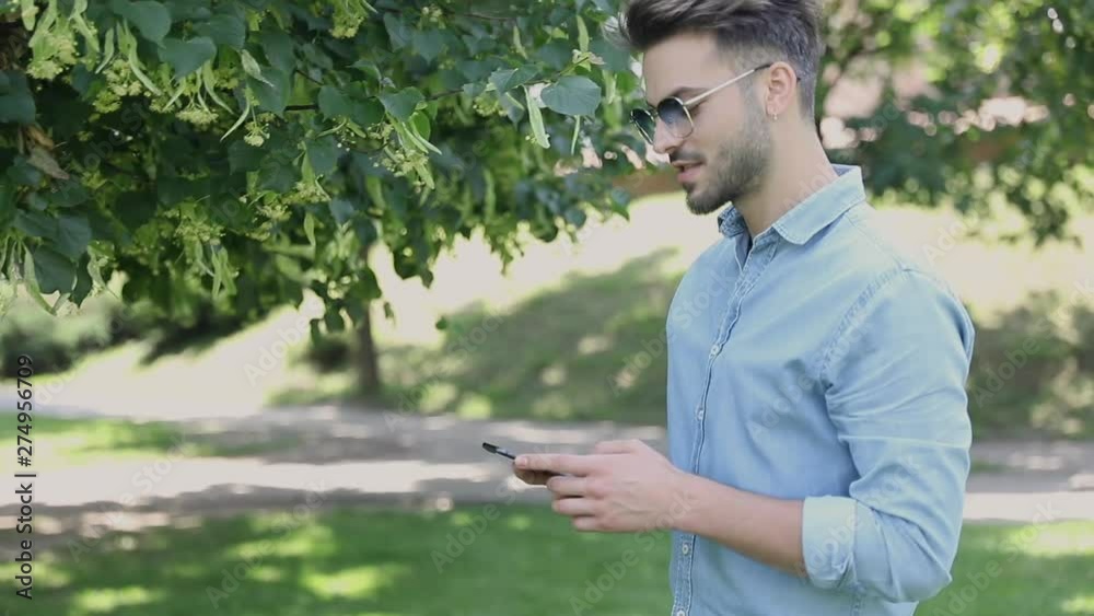 Young casual man wearing sunglasses is walking in the park and messaging on his phone Young casual man wearing sunglasses is walking in the park and messaging on his phone