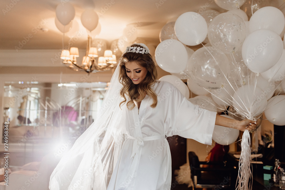 Pretty Young Bride Model Holding Bunch of Balloons. Woman Wearing Gown, Looking Down and Keeping in Hand Helium Air Bubbles. Colorful Photo of Wedding Day. Hotel Apartment Interior on Background - obrazy, fototapety, plakaty 