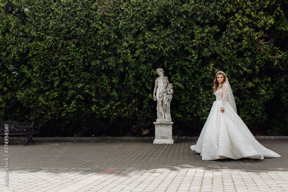 Beautiful Gorgeous Character Bride Walking in Park. Happy Smiling Woman in Wedding Dress Going along Green Trees near Statue and Bench. Young Attractive Caucasian Brunette Model Trendy Marriage Style