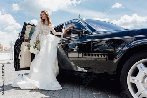 Stylish Bride with Flower Bouquet near Limousine. Pretty Young Woman Dressed White Wedding Dress and Jewelry Diadem with Veil. Girl Holding Roses Bunch and Standing near Black Car. Outside Photo © oksana_bondar