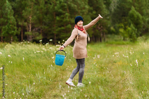 a smiling, happy retired woman goes through the woods with a bucket