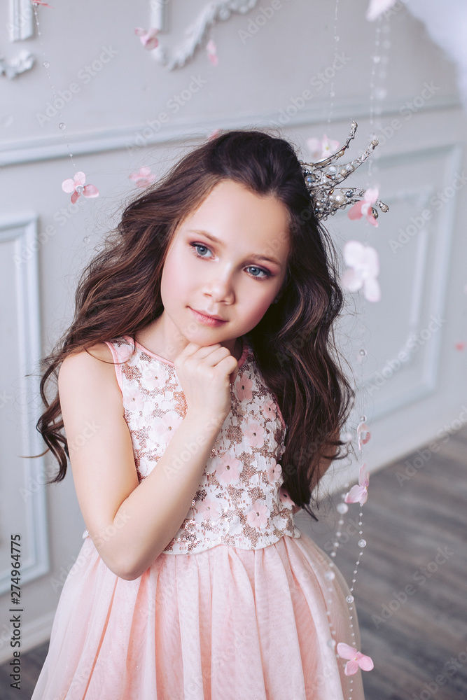 Cute little girl with a dreamy facial expression posing against a white wall in a beautiful pink dress and a crown on her head.