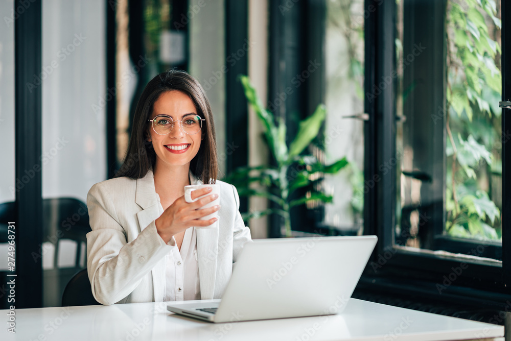 Portrait of elegant young female leader sitting at desk with laptop and holding a mug. Portrait of elegant young female leader sitting at desk with laptop and holding a mug.