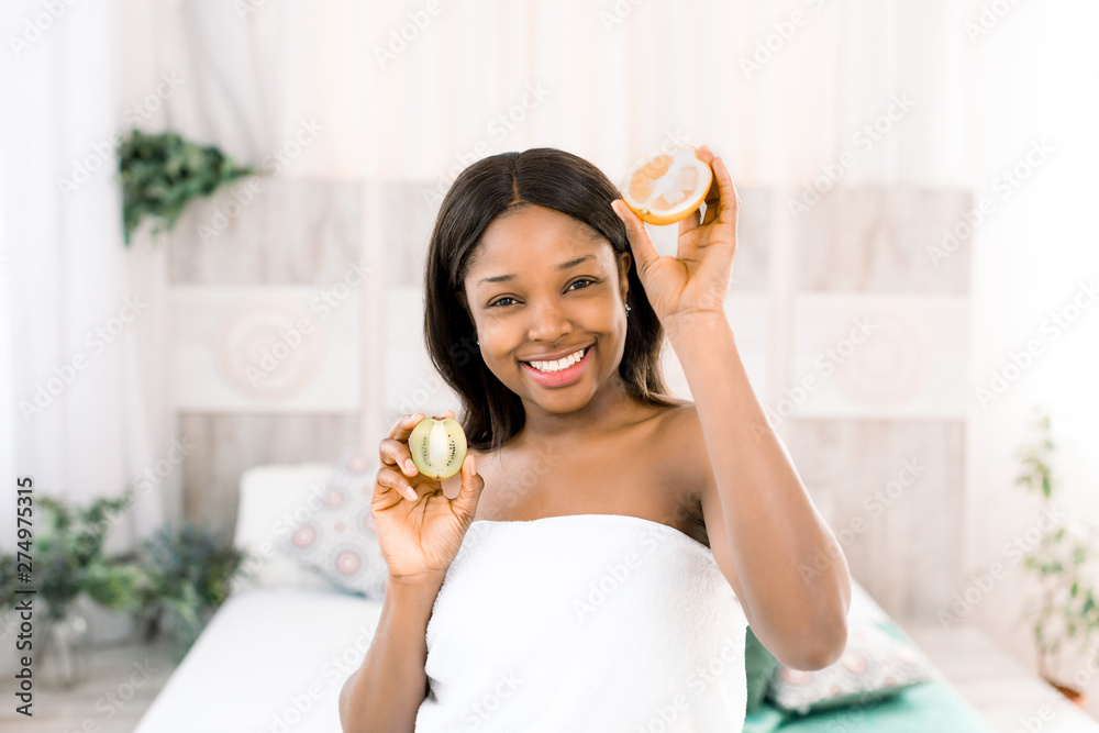 Funny amusing african american young woman holding kiwi and orange halves in hands over light background in spa Funny amusing african american young woman holding kiwi and orange halves in hands over light background in spa