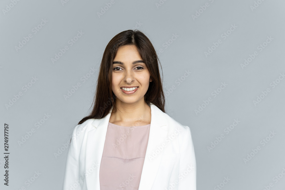 Smiling indian young businesswoman wear white suit looking at camera isolated on grey studio background, happy attractive confident hindu business lady professional head shot close up portrait