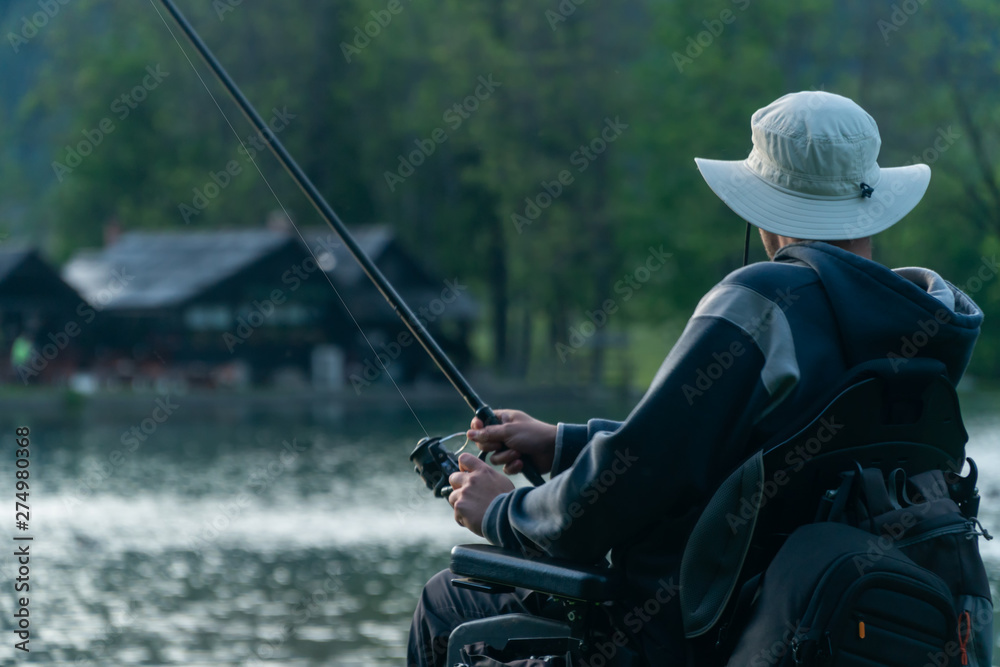 Obraz premium young man in a wheelchair fishing at the beautiful lake in sunset, dawn