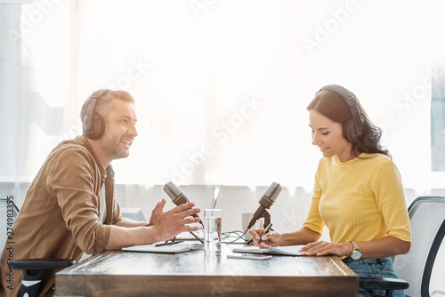 two smiling radio hosts in headphones talking while sitting at table in studio