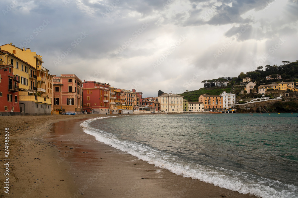 Scenic view of the Bay of Silence in the old fishing village of Sestri Levante with the typical colored houses leaned out on the beach with sun rays filtering through clouds, Liguria, Italy