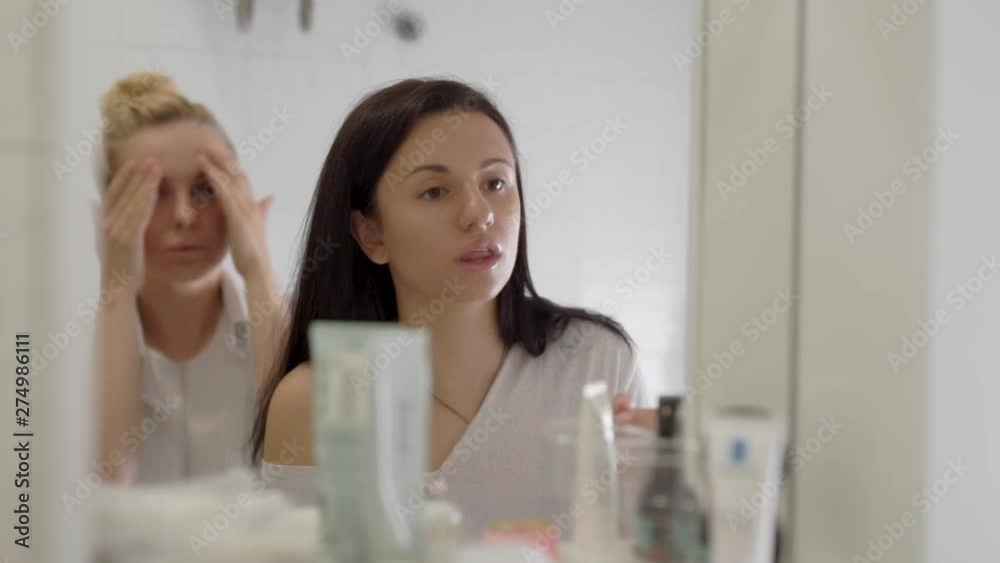 blonde and brunette women are preening in front of mirror in room in morning time together, applying cream and painting lips blonde and brunette women are preening in front of mirror in room in morning time together, applying cream and painting lips