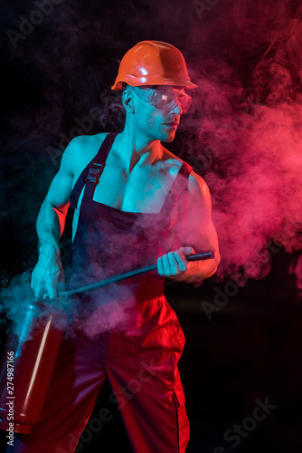 sexy shirtless fireman in overall holding fire extinguisher in smoke on black
