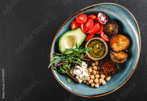 Healthy Buddha bowl dish with avocado, tomato, cheese, chickpea, fresh arugula salad, baked potatoes and sauce pesto in black background. Dieting food, clean eating, top view