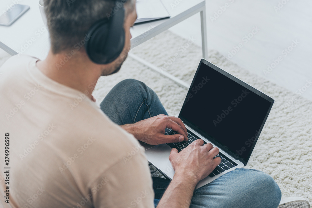 adult man in headphones sitting on floor and using laptop with blank screen adult man in headphones sitting on floor and using laptop with blank screen
