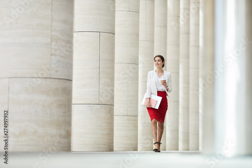 Full length portrait of beautiful business woman wearing red skirt walking towards camera along row of pillars, copy space