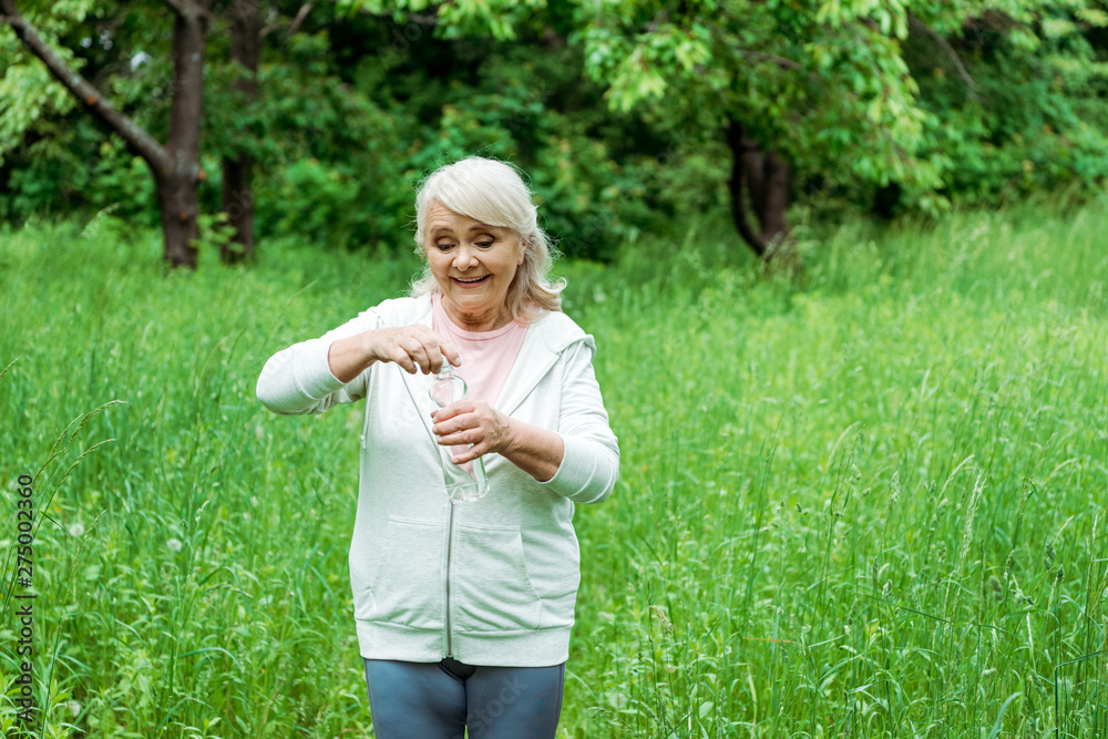 Fototapeta premium cheerful senior woman opening bottle with water in park